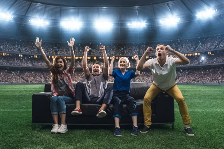 Group of students are watching a soccer moment, sitting on the couch located in the middle of the soccer stadium. Stadium and crowd are made in 3D.